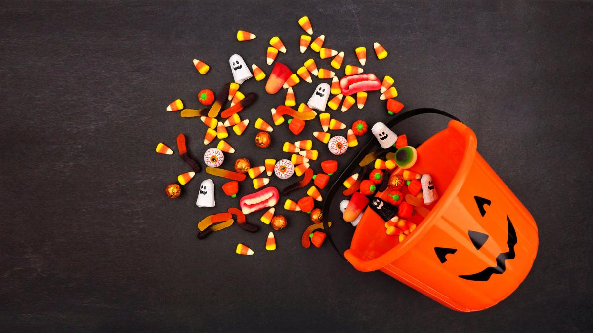 A tipped-over orange jack-o'-lantern bucket with assorted Halloween candies spilling out onto a dark surface.