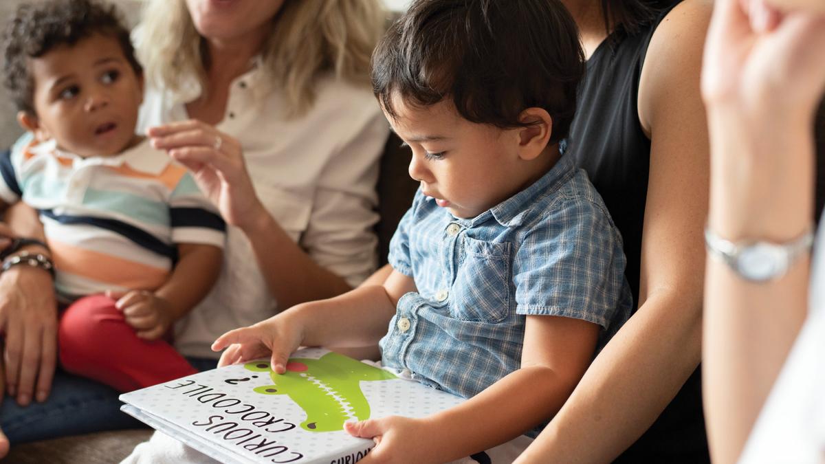 A young child reading a book titled "Raucous Reptiles" while sitting on the lap of an adult, with other adults and a toddler nearby.