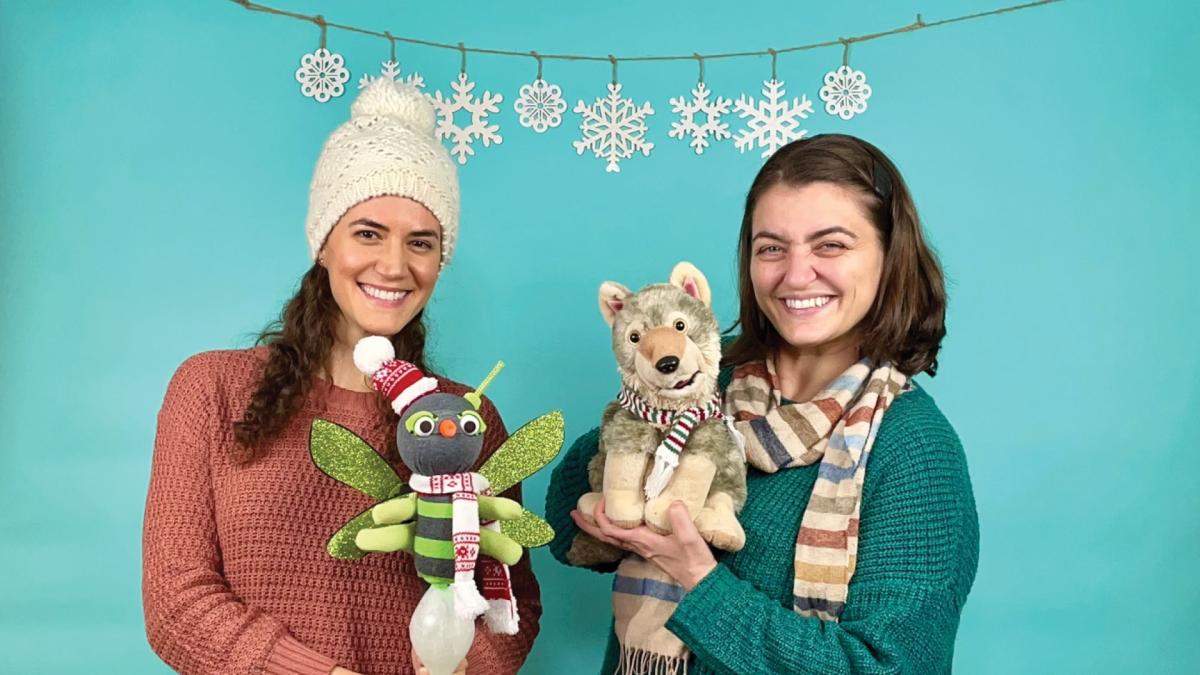 "Two smiling women holding plush toys, with snowflake decorations hanging above them.