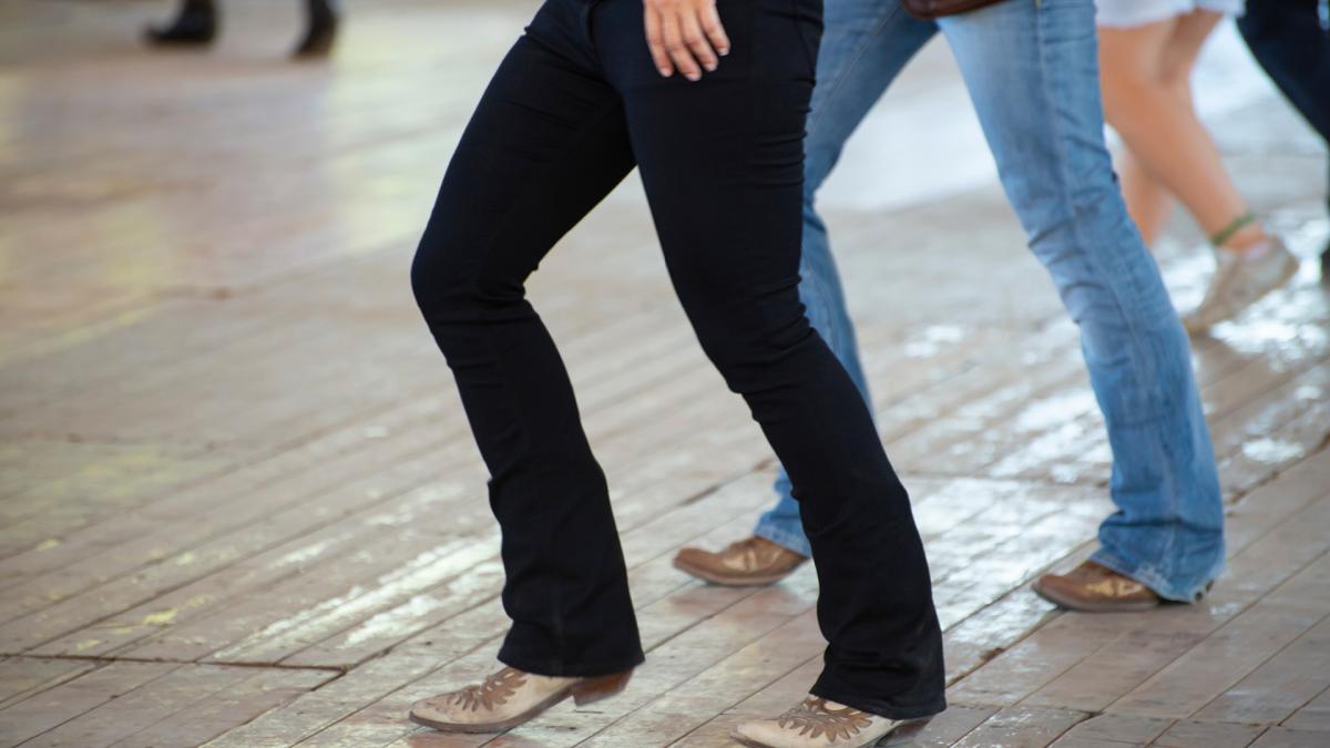 People line dancing indoors with visible lower bodies and casual shoes on a wooden floor.