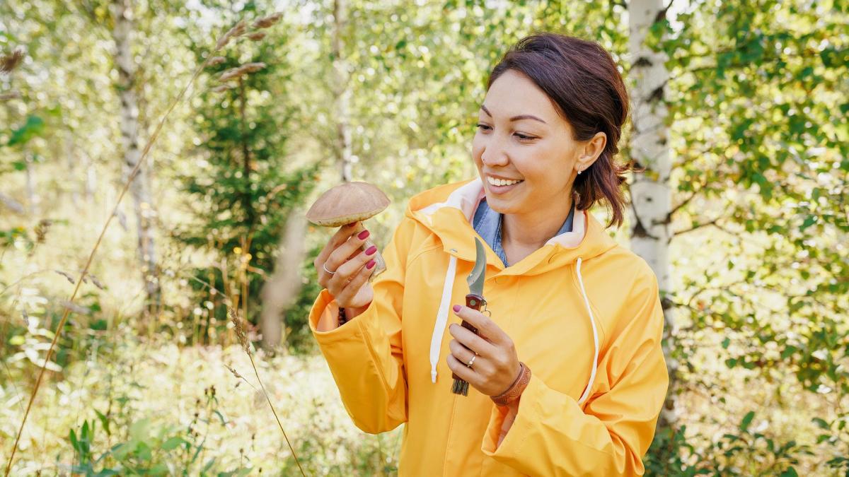 A woman in a yellow jacket holds a mushroom while smiling in a forest.