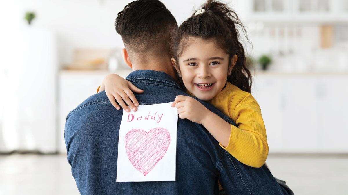 Little girl hugs her dad, holding a drawing with a pink heart and the word "Daddy" on his back.