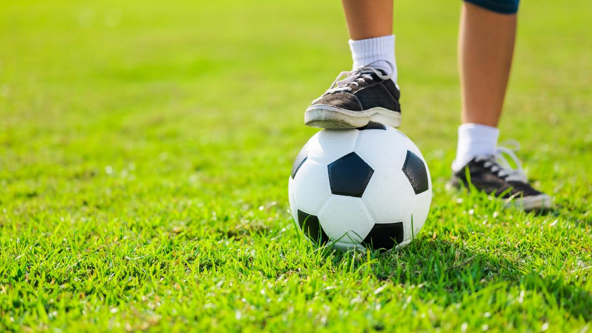 A person in sneakers and socks stands on grass with one foot on a black-and-white soccer ball.