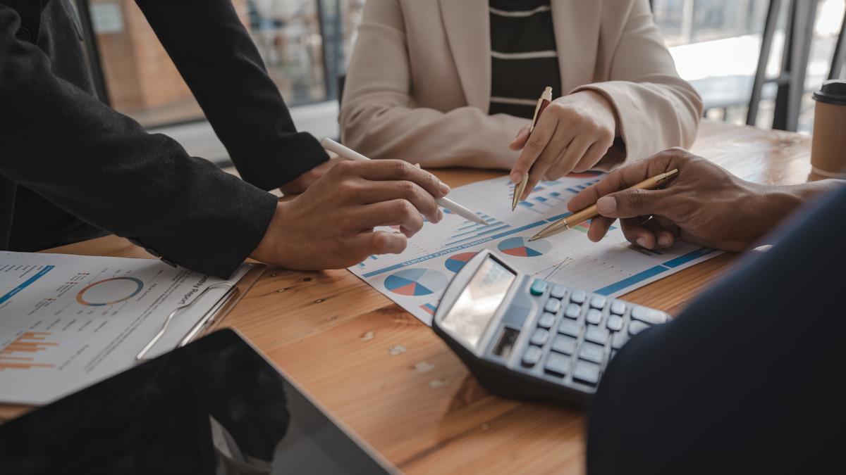 Three people discussing charts and graphs at a table with a calculator and tablet.