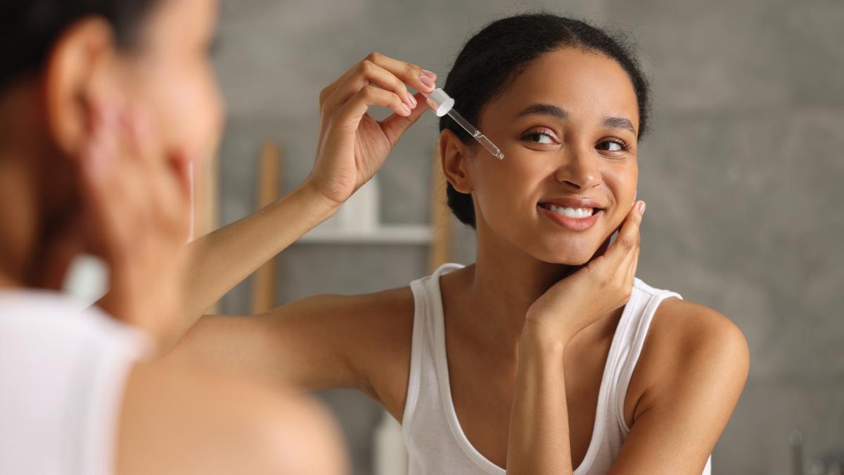 Woman smiling and applying serum to her face with a dropper while looking in the mirror.
