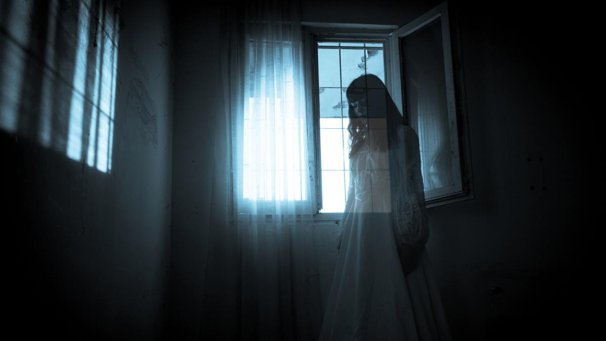 A ghostly figure in a white dress stands by a window with sheer curtains in a dimly lit room.
