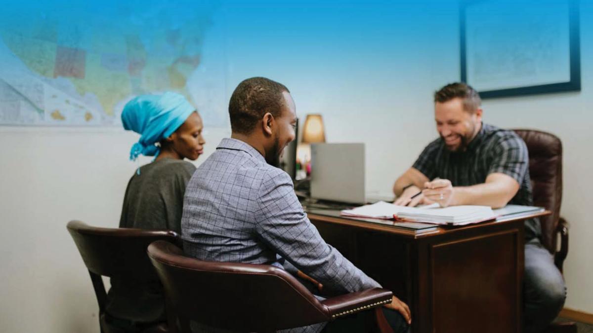 A couple sits across a desk from a smiling man in an office, discussing paperwork.