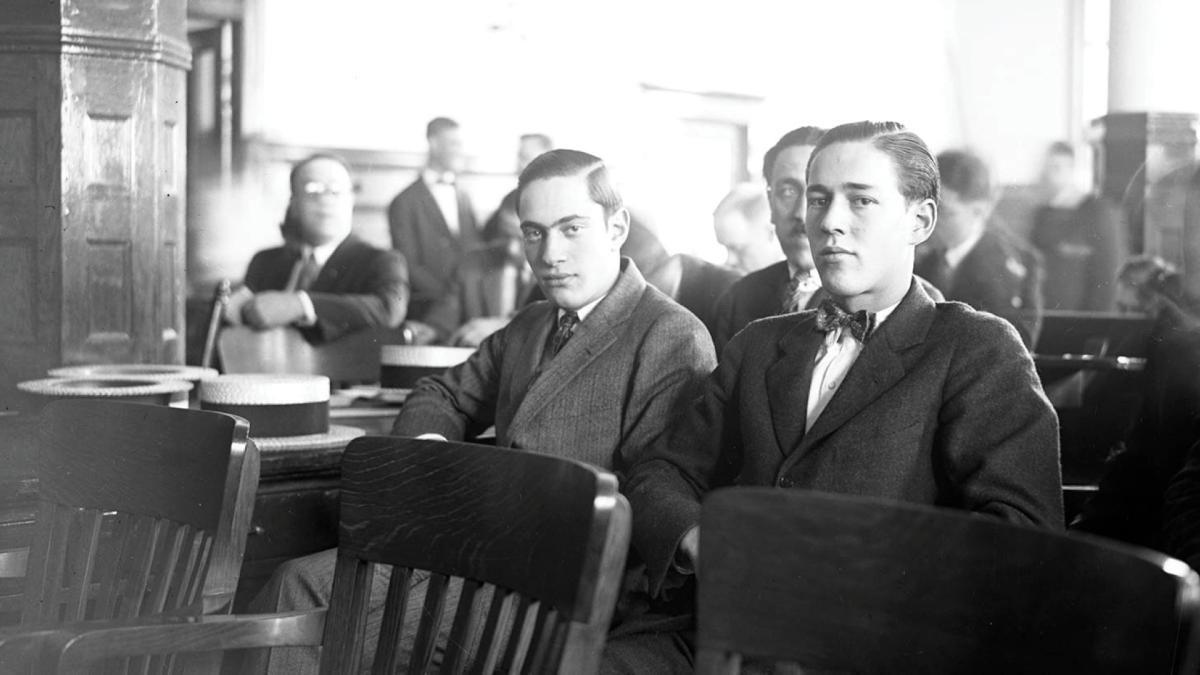 Two young men in suits sit in a courtroom with others visible in the background.
