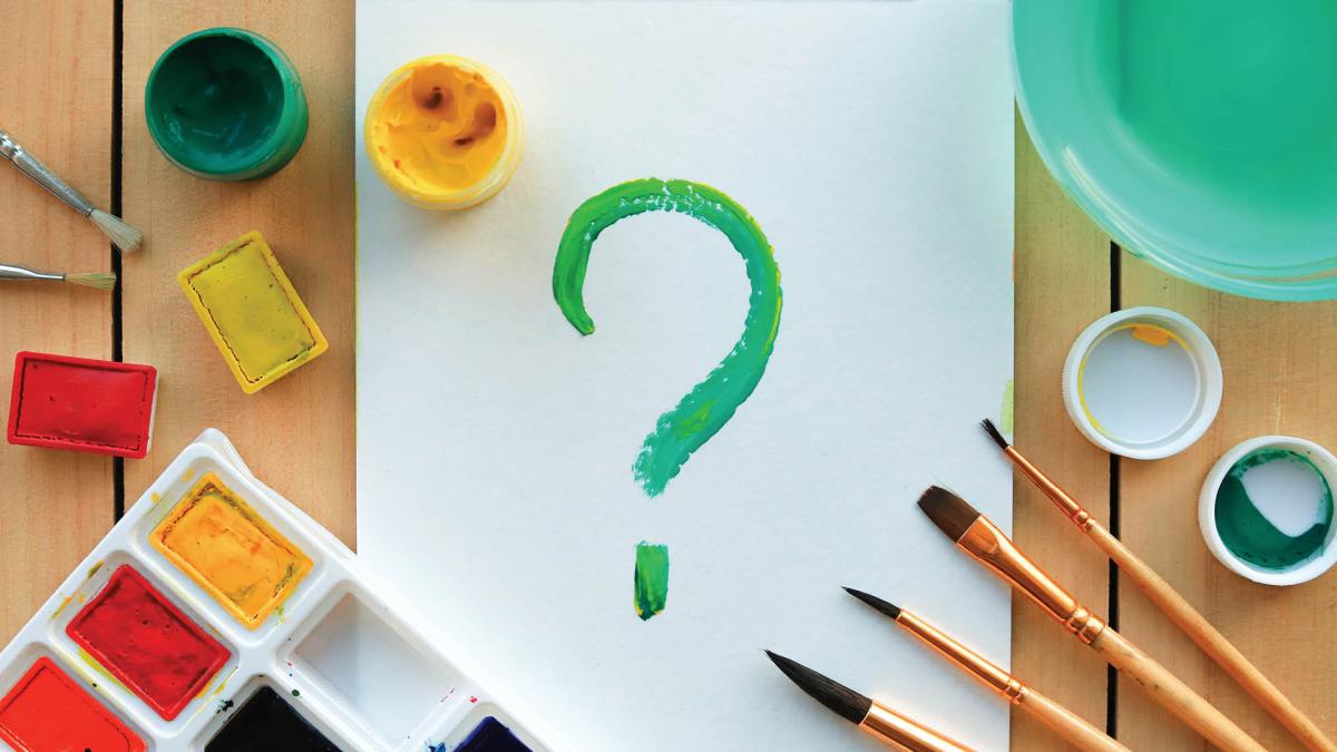 A painted green question mark on paper, surrounded by paint, brushes, and art supplies on a table.