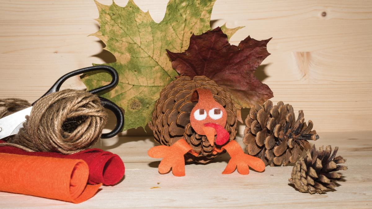A pinecone turkey craft with felt, leaves, twine, scissors, and pinecones on a wooden surface.