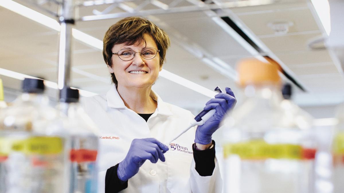 A scientist in a lab coat and gloves holds a pipette, smiling among bottles in a laboratory.